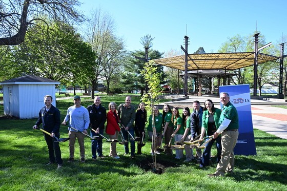 Photo of ceremonial tree planting part of the America250 Tree Planting Ceremony at the Iowa State Fairgrounds. 13 people representing the event's partnering organizations, are holding shovels full of soil before tossing it into the hole to plant the tree.