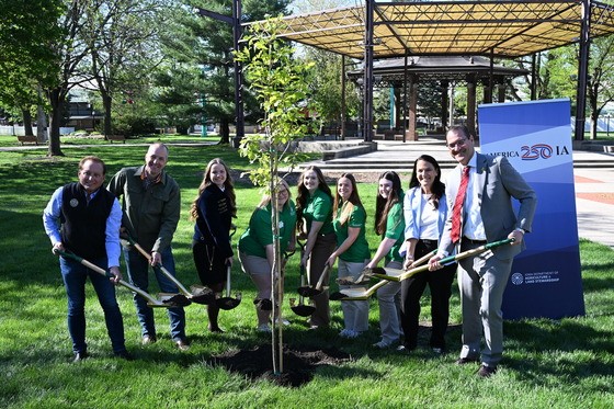Photo of ceremonial tree planting part of the America250 Tree Planting Ceremony at the Iowa State Fairgrounds. 9 people representing the event's partnering organizations and special guests who spoke at the event, are holding shovels full of soil before tossing it into the hole to plant the tree.